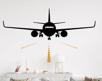 May include: Black silhouette of an aeroplane landing on a white wall with gold landing lights. The aeroplane is centred in the image and is facing the viewer. The wall is plain white and there is a sofa in the foreground.