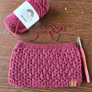 Puede incluir: Un bolso de mano de crochet color burdeos con un patrón de punto de estrella. El bolso tiene una etiqueta de cuero marrón con la palabra "EASI" en ella. También se muestra un ovillo de hilo burdeos y un ganchillo.