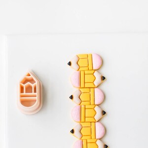 Könnte beinhalten: Sechs gelbe und rosafarbene Fondant-Bleistift-Kekse, vertikal auf einer weißen Oberfläche angeordnet. Links neben den Keksen befindet sich ein rosafarbener Bleistift-Ausstecher.
