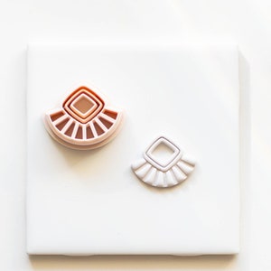 Puede incluir: Dos pares de pendientes geométricos rosa y blanco con una forma de diamante en el centro. Los pendientes están sobre una superficie blanca.