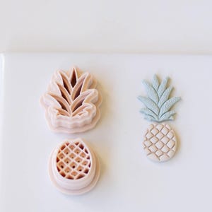 May include: Three pink plastic cookie cutters shaped like pineapples. One cutter has a detailed pineapple design, another has a pineapple leaf design, and the third has a pineapple top design.