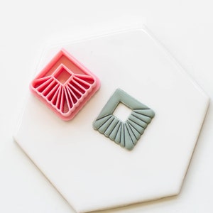 May include: Two cookie cutters, one pink and one gray, shaped like a square with a smaller square cut out in the center. The outer edge of the square has a decorative scalloped design.