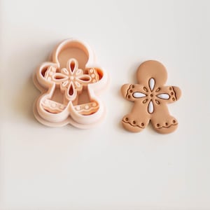May include: A light brown gingerbread man cookie and a matching cookie cutter. The cookie has white accents and a floral design. The cutter is a layered, light pink plastic in the shape of a gingerbread man.