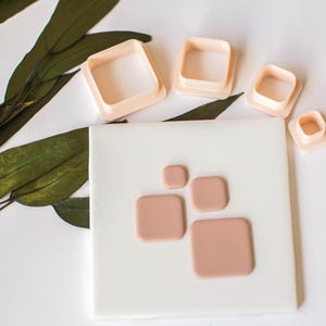 May include: A set of five plastic square cookie cutters in various sizes, with the largest cutter being approximately 2 inches wide. The cutters are on a white surface with a light pink background.