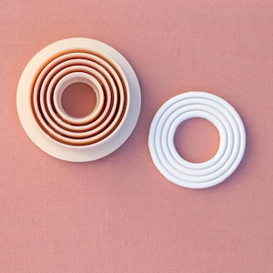 May include: Two sets of round cookie cutters, one set is white and the other is a light beige colour. Both sets have multiple rings that create different sized circles.
