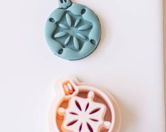 May include: Two Christmas ornament cookie cutters, one is teal blue and the other is pink. Both cutters feature a snowflake design.