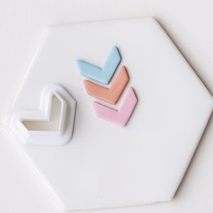 May include: A white plastic arrow-shaped cookie cutter with three clay arrows in blue, pink, and orange.