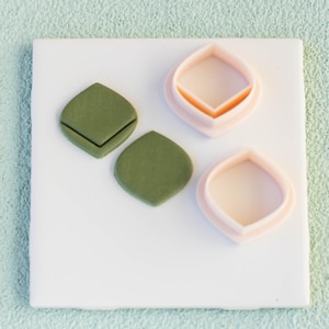 May include: Two green fondant leaf shapes and two pink fondant cookie cutters on a white surface. The cookie cutters are shaped like leaves.