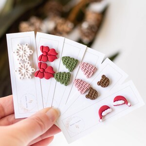 May include: Five cards with pairs of Christmas-themed polymer clay earrings. The earrings are in the shapes of snowflakes, bows, Christmas trees, bears, and Santa hats. The cards are white with a small logo in the bottom right corner.