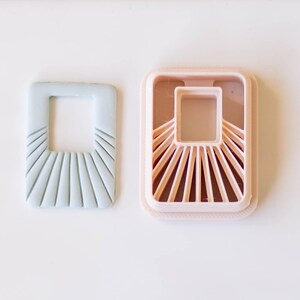 May include: Two square-shaped cookie cutters, one light blue and one light pink, with a sunburst design around a square opening in the center.