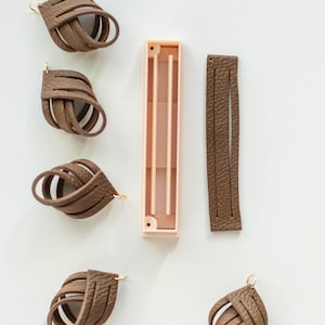May include: Five brown leather earrings with gold jump rings. The earrings are in the shape of a knot. There is a pink plastic mold and a strip of brown leather.