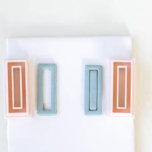 May include: Three rectangular hair clips with a single, narrow, rectangular opening. The clips are in shades of pink, blue, and orange.