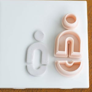May include: A collection of polymer clay cutters in a minimalist design. The cutters are in the shape of a person and a series of arches. The cutters are in shades of light pink and white, and are arranged on a white tile.