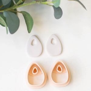 Puede incluir: Dos pares de pendientes de arcilla, un par es rosa claro y el otro par es gris claro. Ambos pares tienen una forma ovalada única con un agujero en el centro.