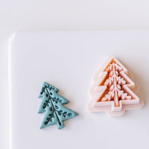 May include: Two Christmas tree-shaped cookie cutters, one is teal blue and the other is light pink.