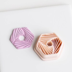 Puede incluir: Dos moldes hexagonales de silicona, uno rosa y otro melocotón, con un diseño geométrico de líneas radiantes.