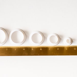 K&ouml;nnte beinhalten: F&uuml;nf runde Ausstecher aus wei&szlig;em Kunststoff in verschiedenen Gr&ouml;&szlig;en, von der gr&ouml;&szlig;ten bis zur kleinsten aufgereiht, mit einem goldenen Lineal darunter. Das Lineal ist in Zoll von 0 bis 12 gekennzeichnet.