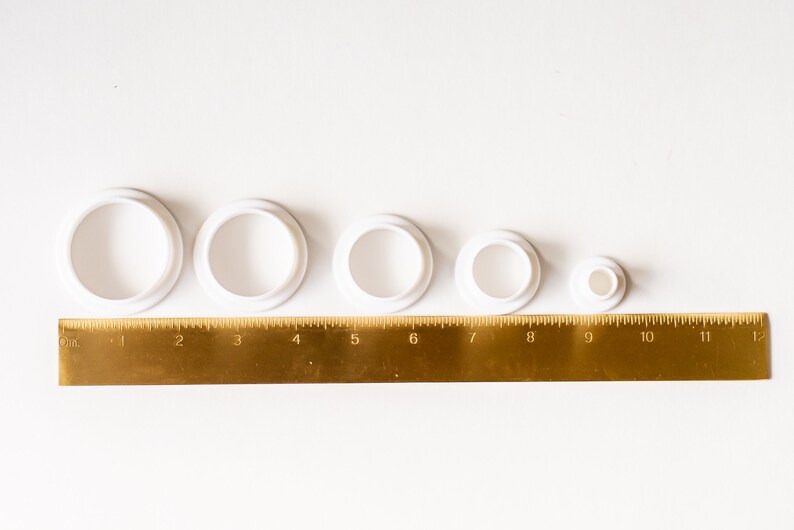 K&ouml;nnte beinhalten: F&uuml;nf runde Ausstecher aus wei&szlig;em Kunststoff in verschiedenen Gr&ouml;&szlig;en, von der gr&ouml;&szlig;ten bis zur kleinsten aufgereiht, mit einem goldenen Lineal darunter. Das Lineal ist in Zoll von 0 bis 12 gekennzeichnet.