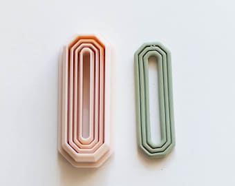May include: Two geometric-shaped cookie cutters. One is a stack of nested, light pink, rectangular shapes. The other is a single, sage green, rectangular shape. Both have a beveled edge and are on a white background.