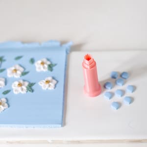 May include: A pink plastic tool for making flower shapes in clay, next to a blue tile with white flowers and leaves, and a group of blue tiles.