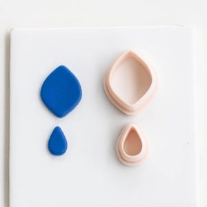 May include: A top-down shot shows two blue clay shapes and two pink clay cutters on a white surface. The blue shapes are a diamond and a teardrop. The cutters are the same shapes. The cutters are made of a light pink plastic.