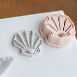 May include: Two seashell-shaped cookie cutters, one gray and one light pink, rest on a white surface. The gray cutter is a finished product, while the pink one is a cutter. The cutters are designed with detailed fan-like ridges.