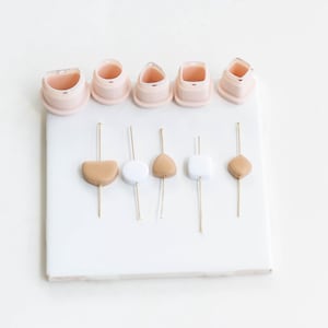 Może przedstawiać: Zestaw wykrawaczy do gliny polimerowej i elementów koralików w różnych kształtach i kolorach. Wykrawacze są jasnoróżowe, a koraliki beżowe i białe. Przedmioty ułożone są na białej powierzchni, idealne do rękodzieła i tworzenia biżuterii.