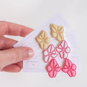 May include: A hand holds three cards displaying colorful floral-shaped earrings. The earrings are in three color schemes: yellow with a cream outline, pink with a white outline, and a darker pink with a white outline. The cards have the text "Blushes + Neutrals".