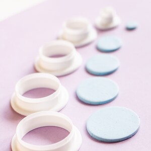 K&ouml;nnte beinhalten: Ein Satz wei&szlig;er Kunststoff-Kreisausstecher und blaue Fondantkreise auf einer violetten Oberfl&auml;che. Die Ausstecher sind in absteigender Reihenfolge angeordnet, wobei der gr&ouml;&szlig;te Ausstecher unten und der kleinste oben liegt. Die Fondantkreise sind &auml;hnlich angeordnet, wobei der gr&ouml;&szlig;te Kreis unten und der kleinste oben liegt.
