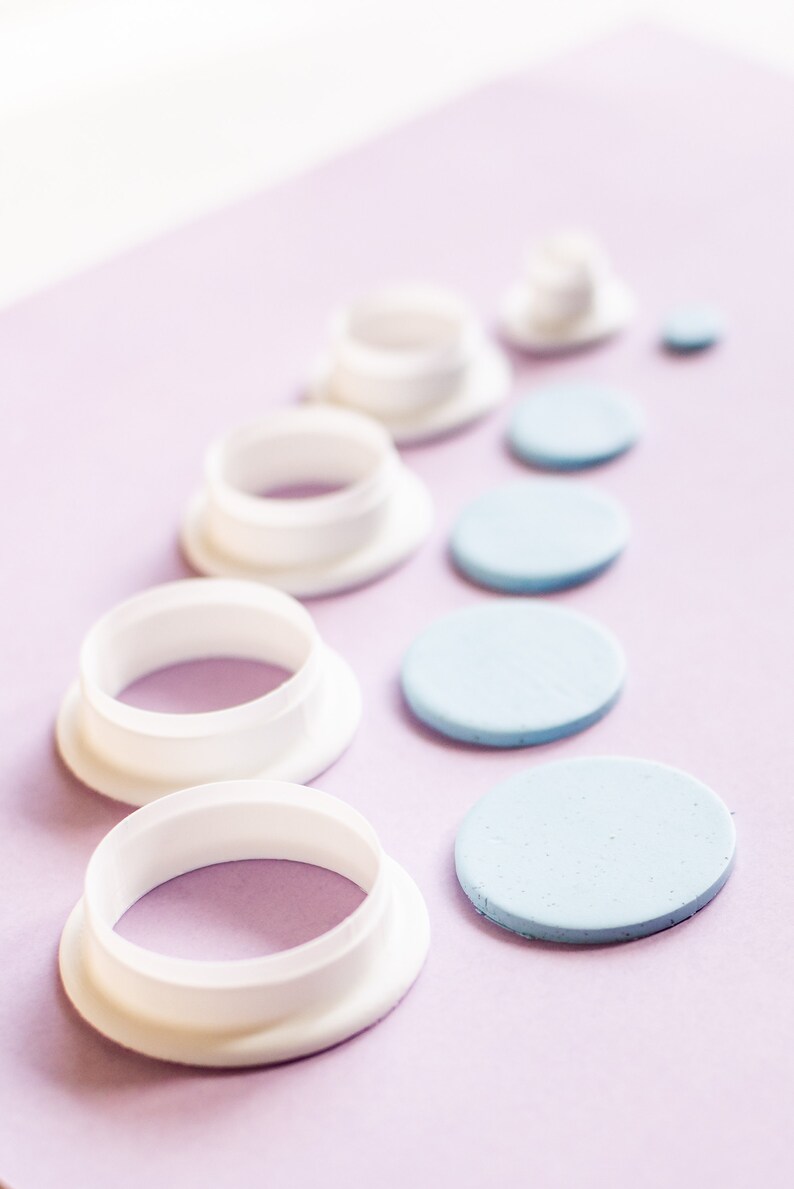 K&ouml;nnte beinhalten: Ein Satz wei&szlig;er Kunststoff-Kreisausstecher und blaue Fondantkreise auf einer violetten Oberfl&auml;che. Die Ausstecher sind in absteigender Reihenfolge angeordnet, wobei der gr&ouml;&szlig;te Ausstecher unten und der kleinste oben liegt. Die Fondantkreise sind &auml;hnlich angeordnet, wobei der gr&ouml;&szlig;te Kreis unten und der kleinste oben liegt.