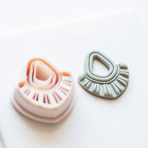 Puede incluir: Dos pendientes de arcilla en forma de abanico. Un pendiente es de color rosa claro con un centro rosa oscuro y naranja. El otro pendiente es de color verde claro con un centro verde oscuro.