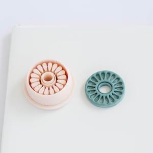 May include: Two floral-shaped polymer clay cutters. One is a light pink color, and the other is teal. The cutters are designed to create flower shapes for crafts or baking. The cutters are on a white surface.