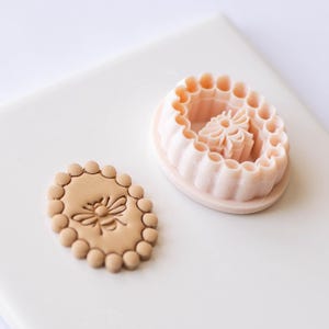 Puede incluir: Un cortador de galletas ovalado rosa claro con un diseño floral y una galleta en relieve a juego. El cortador tiene un borde festoneado y un centro floral detallado. La galleta en relieve presenta un diseño de abeja.