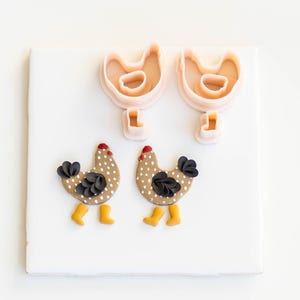 Puede incluir: Dos pollos de arcilla marrón y blanco con lunares, alas negras y patas amarillas. Dos cortadores de galletas de pollo de plástico rosa.