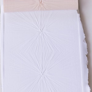 May include: A close-up of white paper with a textured, starburst-like pattern. A rolled piece of light pink paper sits above the patterned paper. The edges of the paper are uneven, giving it a handmade appearance. The image is well-lit.