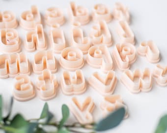 May include: A set of plastic alphabet cookie cutters in a light peach colour. The cutters are arranged in a random pattern on a white surface.