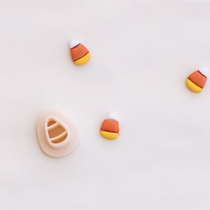 Könnte beinhalten: Fünf orange, gelbe und weiße Candy-Corn-Formen und ein weißer Plastik-Candy-Corn-Ausstecher auf weißem Hintergrund.