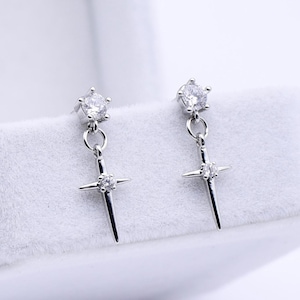 May include: A pair of silver earrings with a dangling cross design, each featuring a clear, round gemstone at the top and a smaller gemstone at the center of the cross. The earrings are displayed in an open white jewelry box.
