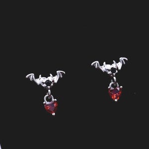 May include: A pair of silver-toned stud earrings shaped like bats with outstretched wings, each featuring a small, dangling red heart-shaped gemstone. The earrings are presented inside a square white box with a black interior lining.
