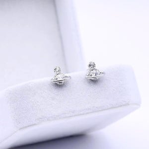 May include: A pair of silver-toned stud earrings shaped like a planet with a surrounding ring, featuring small clear crystals embedded in the design. The earrings are displayed resting on the velvet-lined interior of a white jewelry box.
