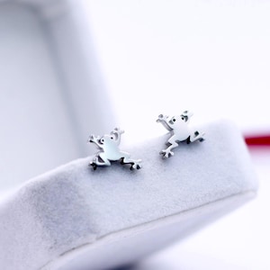 May include: A pair of silver frog stud earrings displayed in an open white velvet jewelry box. The earrings are small, detailed, and feature a frog design. A red ribbon is visible in the background, adding a pop of color to the scene.
