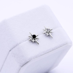 May include: A pair of starburst stud earrings, one with a black center stone and the other with clear crystals. The earrings are displayed on a white, hexagonal jewelry box with a soft, velvety texture.