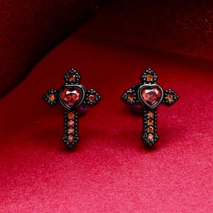 May include: A pair of black metal stud earrings shaped like crosses. Each earring features a central heart-shaped red gemstone surrounded by smaller, round orange-red accent stones embedded along the arms of the cross design.