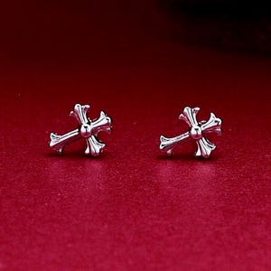May include: A pair of silver-toned stud earrings shaped like ornate crosses. Each earring features a central circular detail with four flared arms extending outward, creating a symmetrical design with a polished metallic finish and reflective surface.