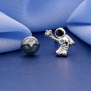May include: A pair of silver-toned stud earrings featuring an astronaut figure with a black visor and a small round planet charm with a central clear stone. The astronaut is depicted in a floating pose, while the planet has a textured surface.