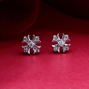 May include: A pair of silver-toned stud earrings featuring a four-pointed cross design. Each earring is set with small, clear, sparkling stones along the arms and a single, larger, clear stone at the center of the cross-shaped metal frame.