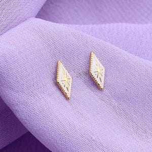 May include: A pair of small, gold-toned stud earrings shaped like elongated diamonds. Each earring features a textured surface with a central starburst engraving and a delicate beaded border, providing a refined and detailed appearance for these jewelry pieces.