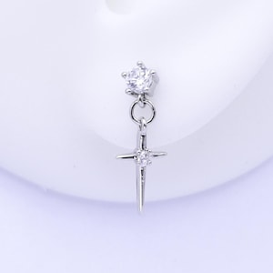 May include: A silver earring with a dangling cross design. The earring features a small, round, clear gemstone at the top and a smaller gemstone in the center of the cross. The earring is displayed next to a white ear.