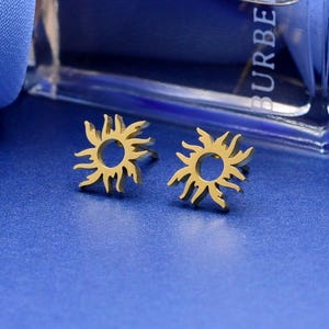May include: A pair of gold-toned stud earrings featuring a sunburst design with a circular center and radiating flame-like points. The earrings are positioned in front of a clear glass bottle with the text BURBERRY visible on the side.