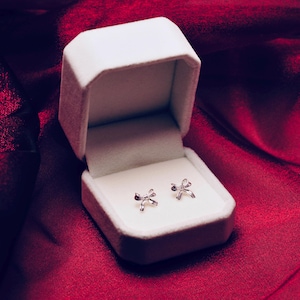 Puede incluir: Un par de pendientes de bot&oacute;n plateados con forma de lazo expuestos en una caja de joyer&iacute;a blanca abierta. La caja descansa sobre una tela de sat&eacute;n rojo intenso, creando una presentaci&oacute;n lujosa. Los pendientes son peque&ntilde;os y delicados.